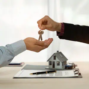 Person handing keys to another, with a house model, documents, and pen on the table, symbolizing a real estate deal - Kollias Law